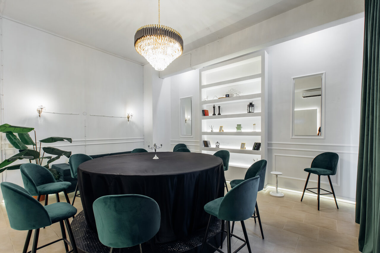 Арендовать Rubin Loft (Рубин Лофт). Зал Империал. Просторная комната с круглым столом, покрытым черной скатертью, окружена зелеными стульями. На стене белые полки с декором. Светильник с хрустальной люстрой по центру. Светлые стены, паркетный пол, зелёное растение слева.
