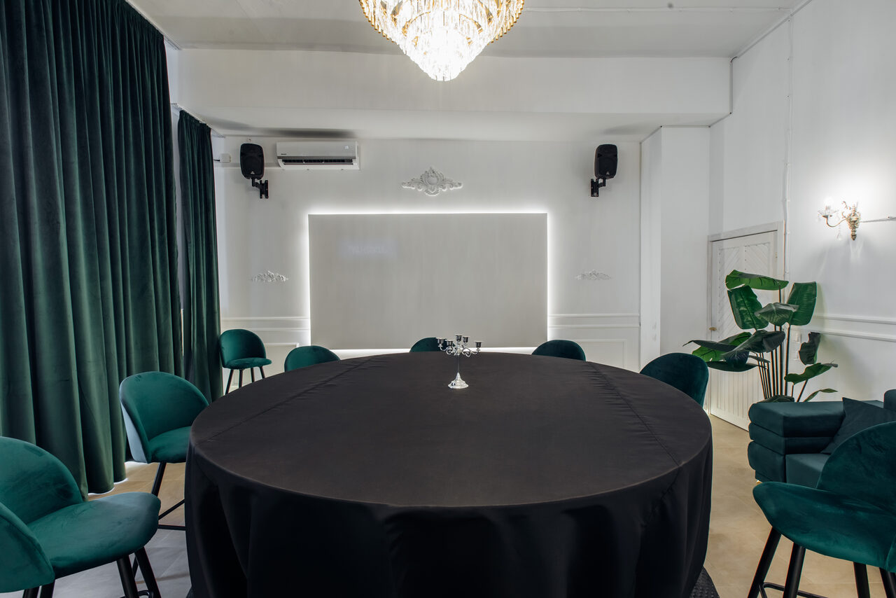 Арендовать Rubin Loft (Рубин Лофт). Зал Империал. Просторная комната с круглым столом, покрытым черной скатертью. Вокруг расположены стулья с зелеными бархатными обивками. На стене белая панель с подсветкой, темные шторы, два настенных светильника и кондиционер. Пленочное растение в углу.