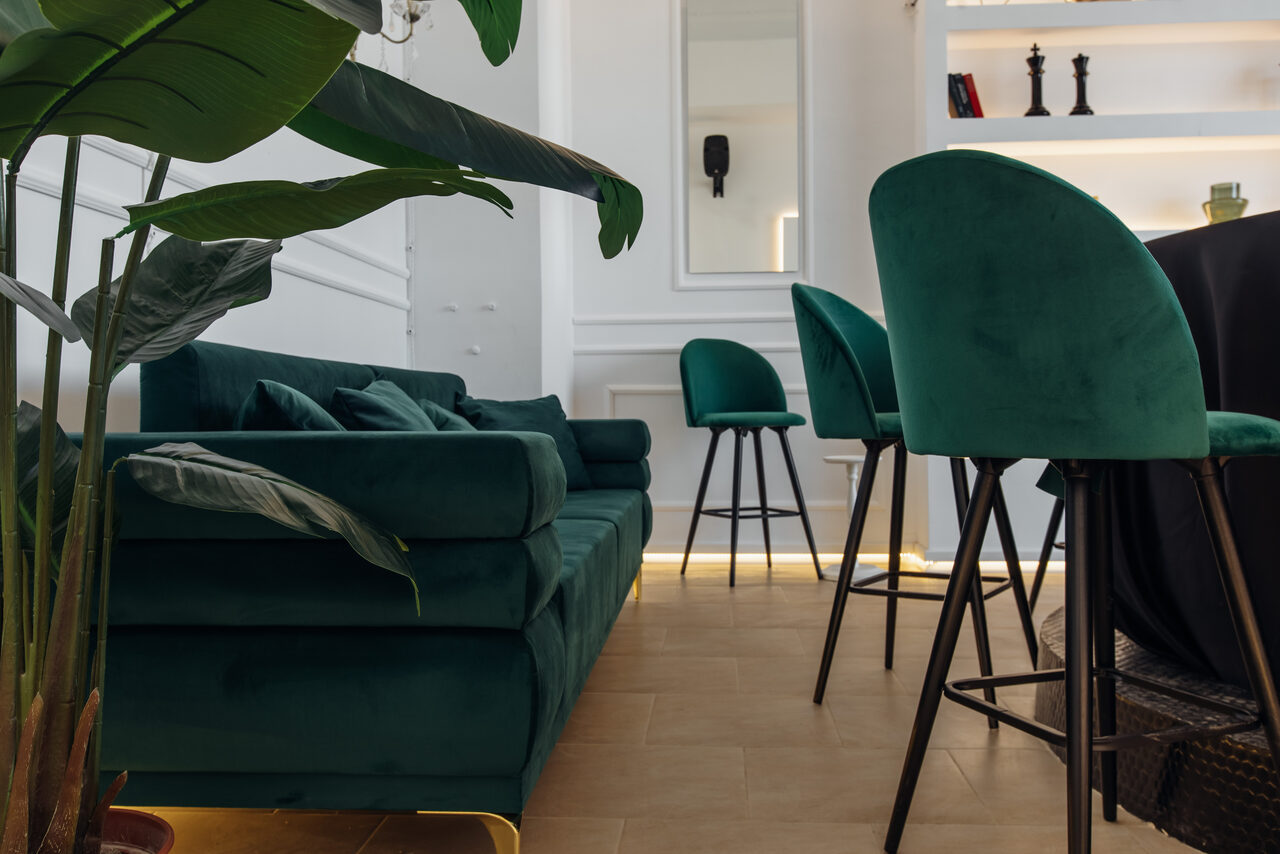 Арендовать Rubin Loft (Рубин Лофт). Зал Империал. Гостиная с деревянным полом, глубокие зеленые диваны и стулья, белые стены, подсвеченные полки с декором, зеркало на стене, зелёные комнатные растения, хорошее естественное освещение.