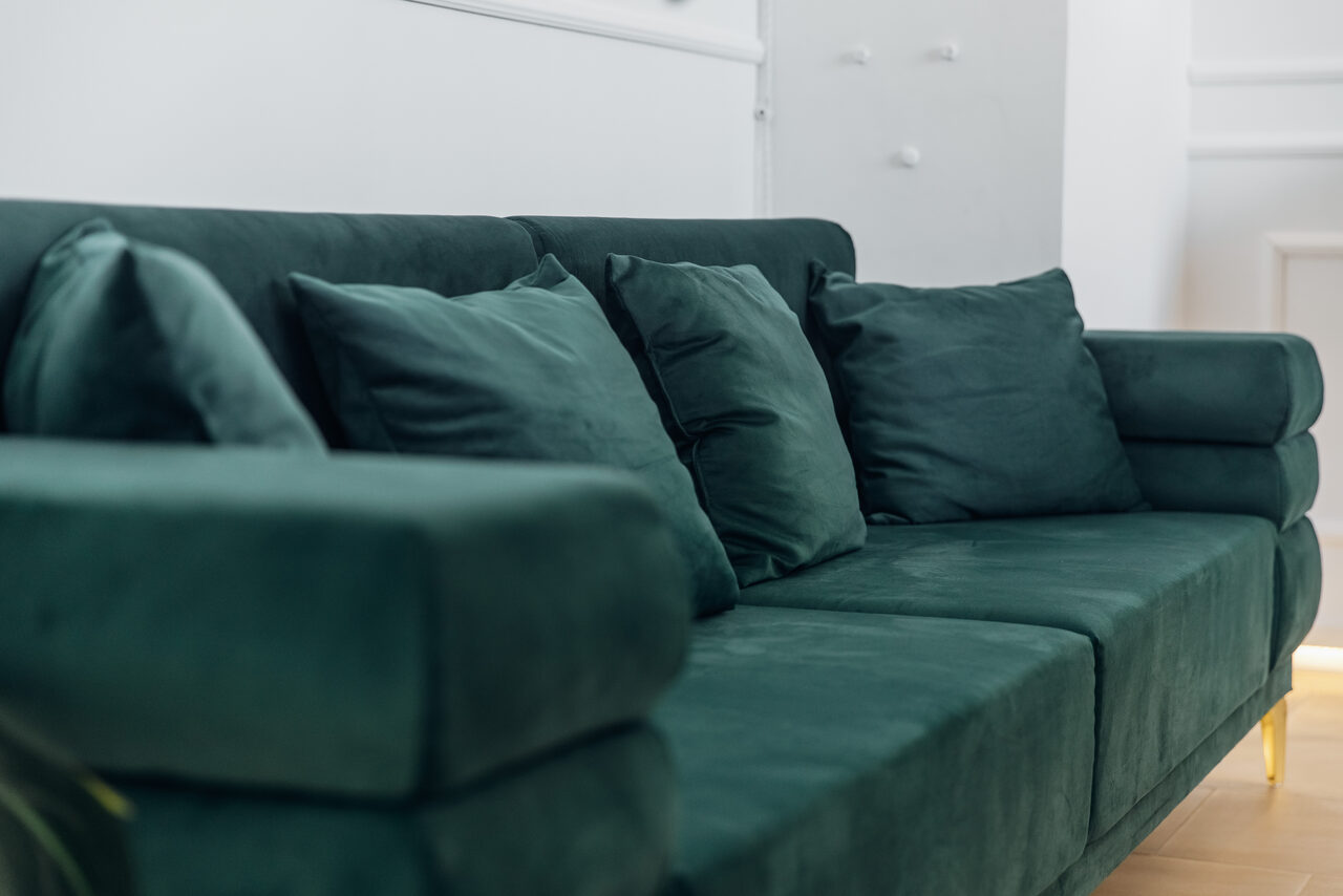 Арендовать Rubin Loft (Рубин Лофт). Зал Империал. Зеленый бархатный диван с несколькими подушками в минималистичном интерьере. Светлые стены, светлое деревянное напольное покрытие. Элементы декора белого цвета на стене.