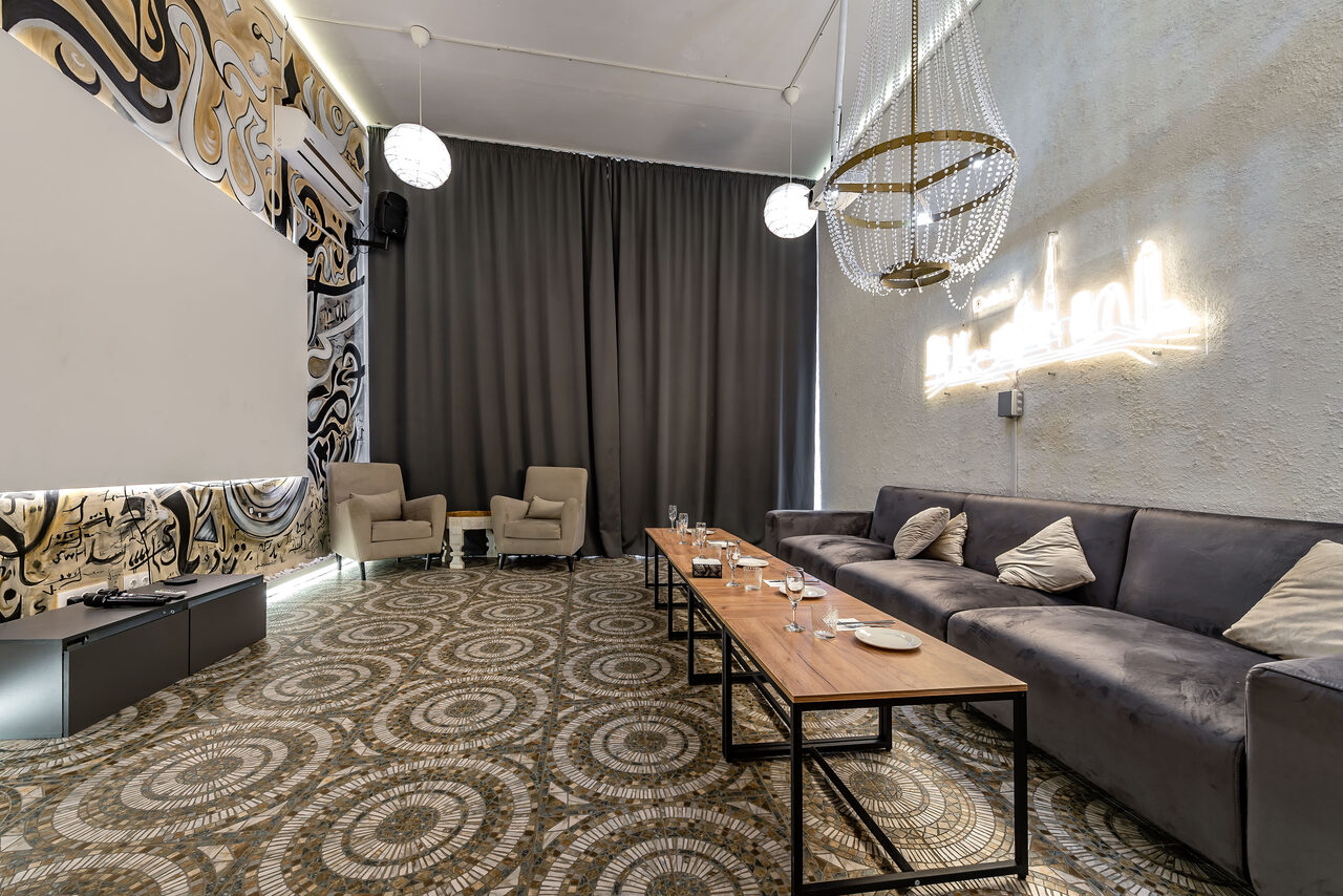 Арендовать Rubin Loft (Рубин Лофт). Зал Дубай. Просторная комната с диваном, креслами и длинным деревянным столом. На полу геометрический узор, стены с абстрактным рисунком и нейтрального цвета. Подвесные светильники и неоновая надпись на стене. Тёмные шторы закрывают окна.