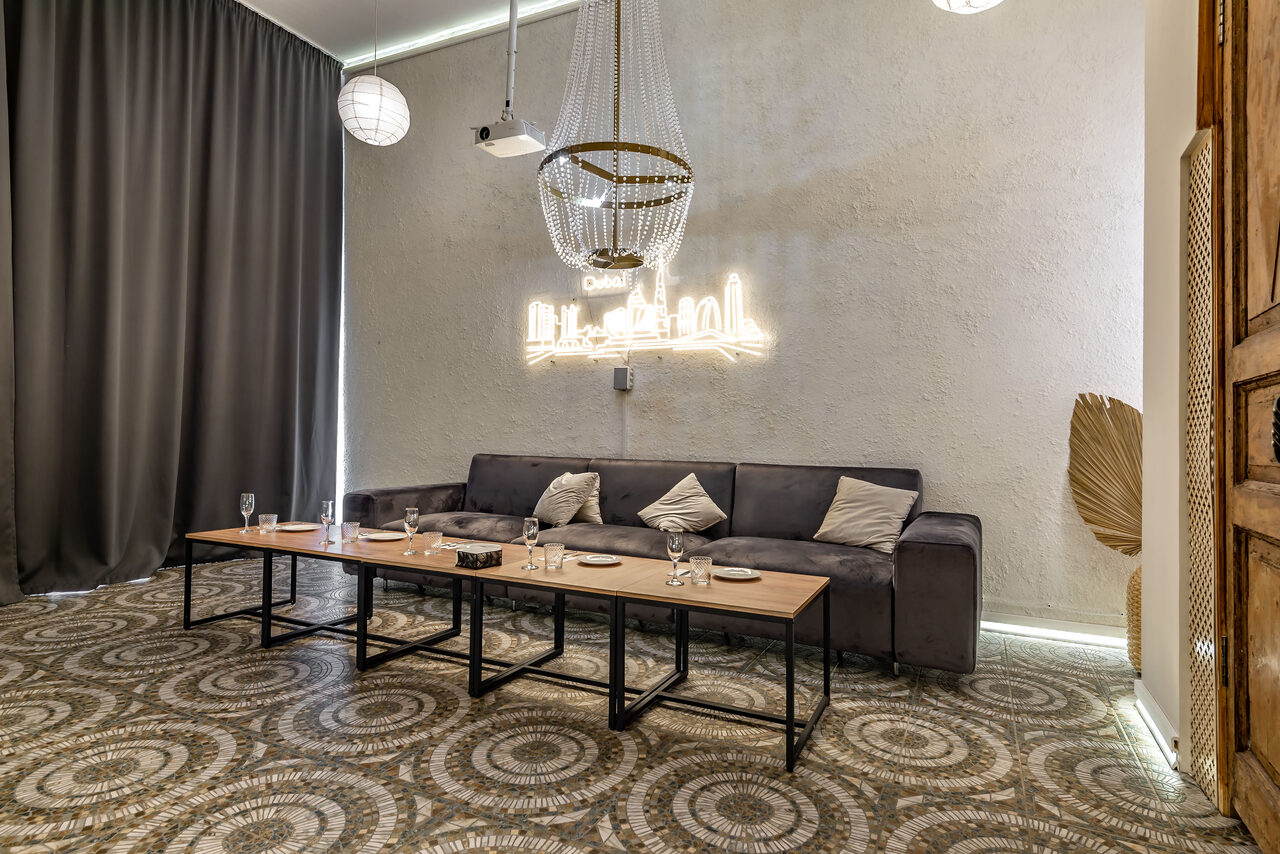 Арендовать Rubin Loft (Рубин Лофт). Зал Дубай. Просторная гостиная с темным диваном, перед которым расположен длинный деревянный стол на металлических опорах. Стены светлые, пол с узорчатым покрытием. Потолок украшает большая люстра и неоновая панель. Справа висят темные шторы.
