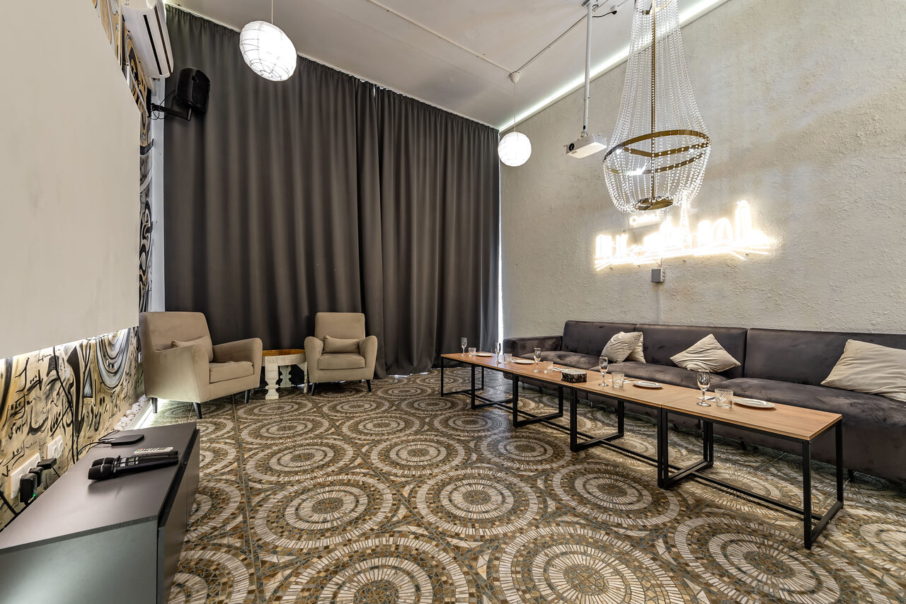 Арендовать Rubin Loft (Рубин Лофт). Зал Дубай. Комната с диваном, креслами и длинным столом; гладкие серые стены и занавески; подвесные светильники; декоративный пол с узором; неоновая надпись на стене; элементы декора и аудиооборудование.