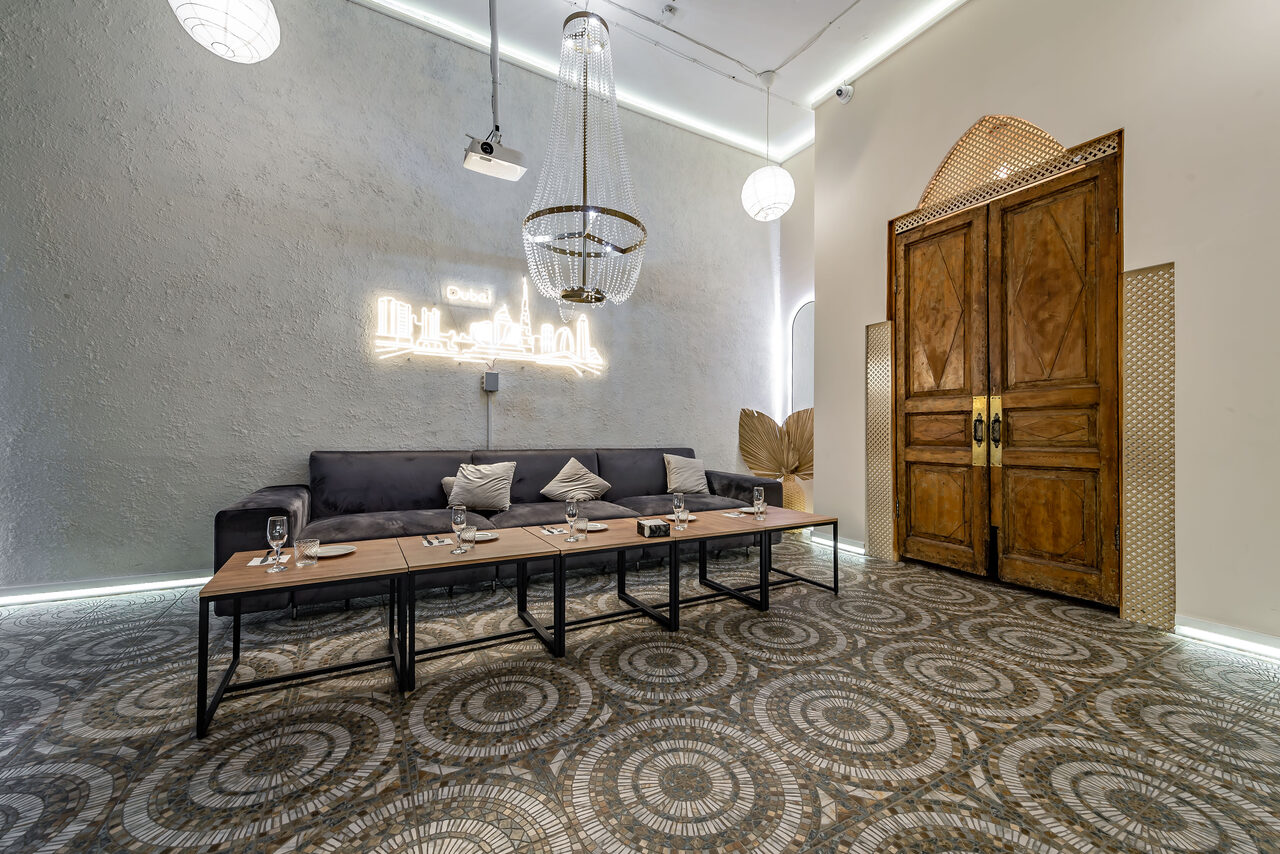 Арендовать Rubin Loft (Рубин Лофт). Зал Дубай. Лофт-пространство с серым диваном и журнальными столиками, серо-коричневые узорчатые плиточные полы, деревянные двустворчатые двери, декоративные светильники и неоновая надпись на стене.