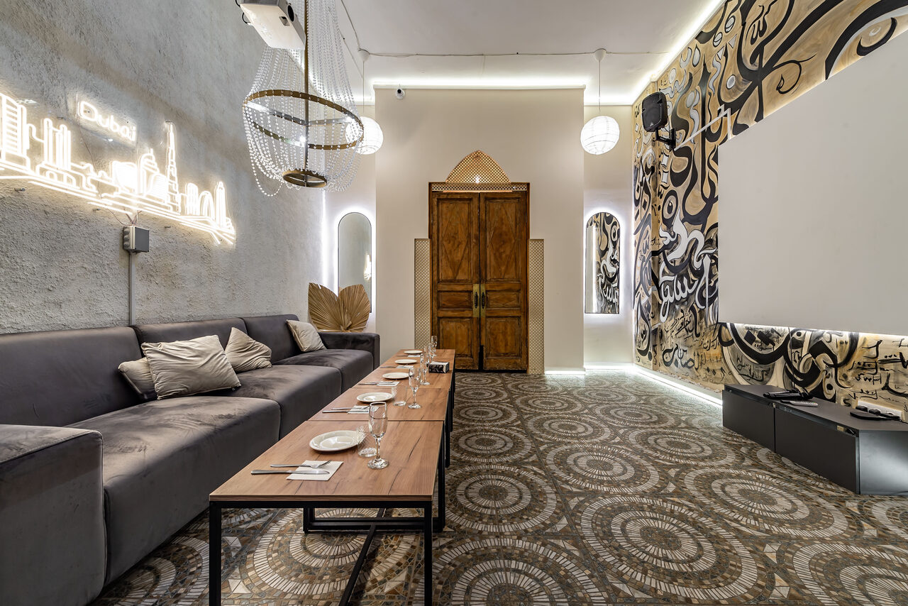 Арендовать Rubin Loft (Рубин Лофт). Зал Дубай. Просторное помещение с мягким диваном и деревянным столом, сервированным тарелками и стаканами. Светлая текстура стен с граффити, декоративные светильники и неоновая надпись. Декоративные двери, геометрическая плитка на полу.