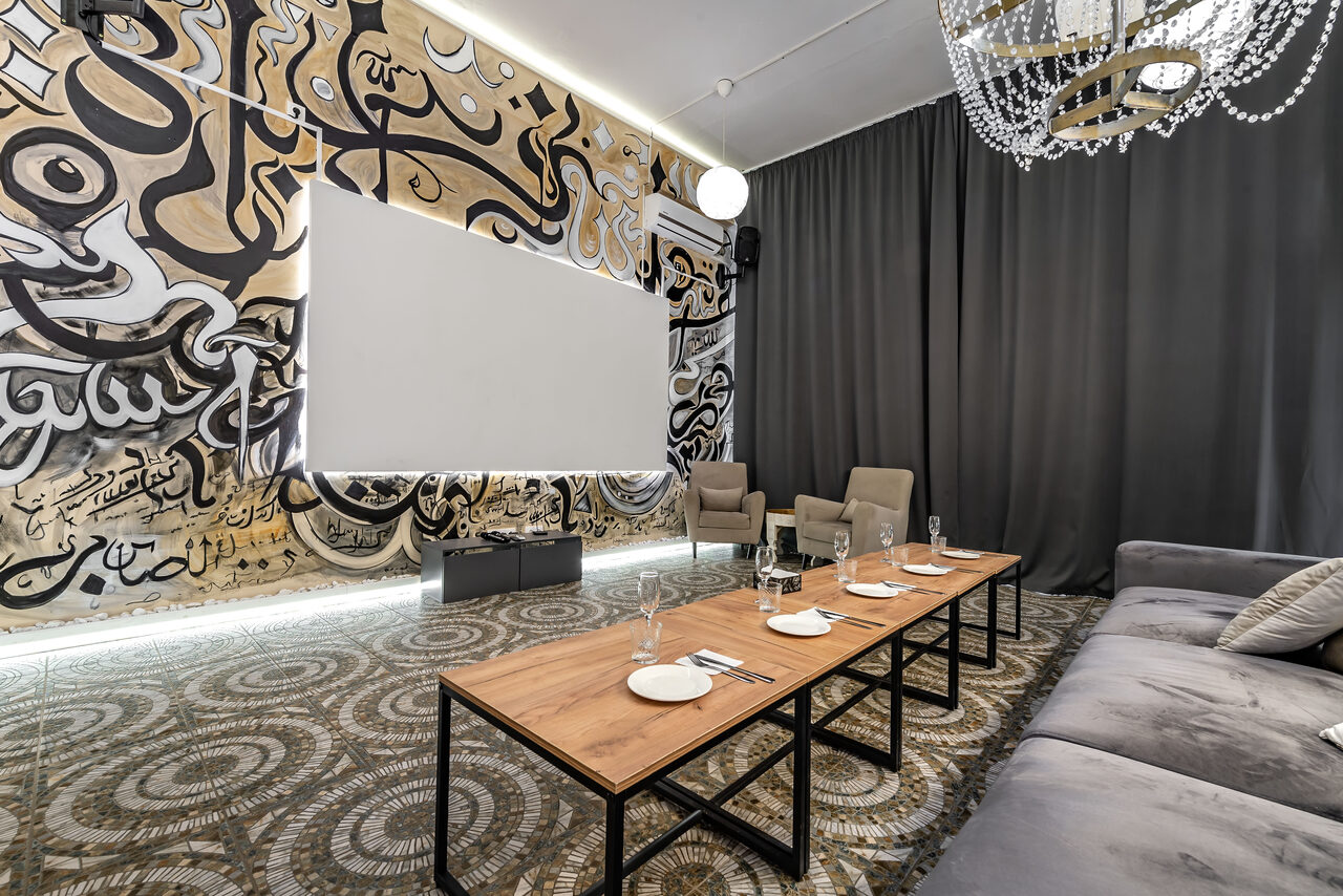 Арендовать Rubin Loft (Рубин Лофт). Зал Дубай. Просторное помещение с проектором и экраном, стильные граффити на стенах, серые шторы. Удобные кресла, длинный деревянный стол с посудой, мягкий диван. Потолочная люстра с украшениями, светлое декоративное освещение, узорчатый ковролин.