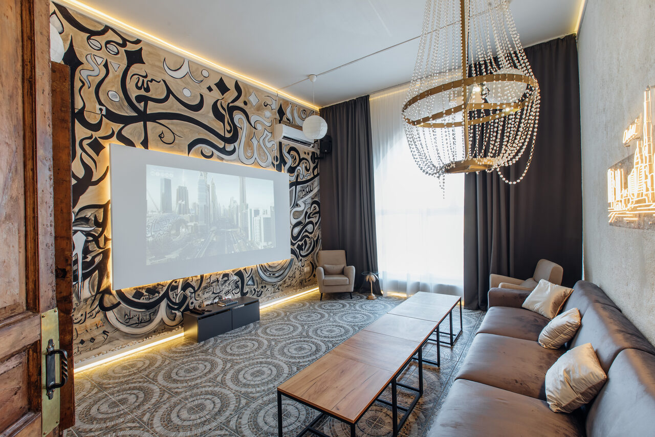 Арендовать Rubin Loft (Рубин Лофт). Зал Дубай. Комната с проектором и экраном, декоративные узоры на стенах, большое окно с темными занавесками, мягкий диван и кресла, длинные деревянные столы, ковровое покрытие, крупная люстра, декоративные светильники на стене.