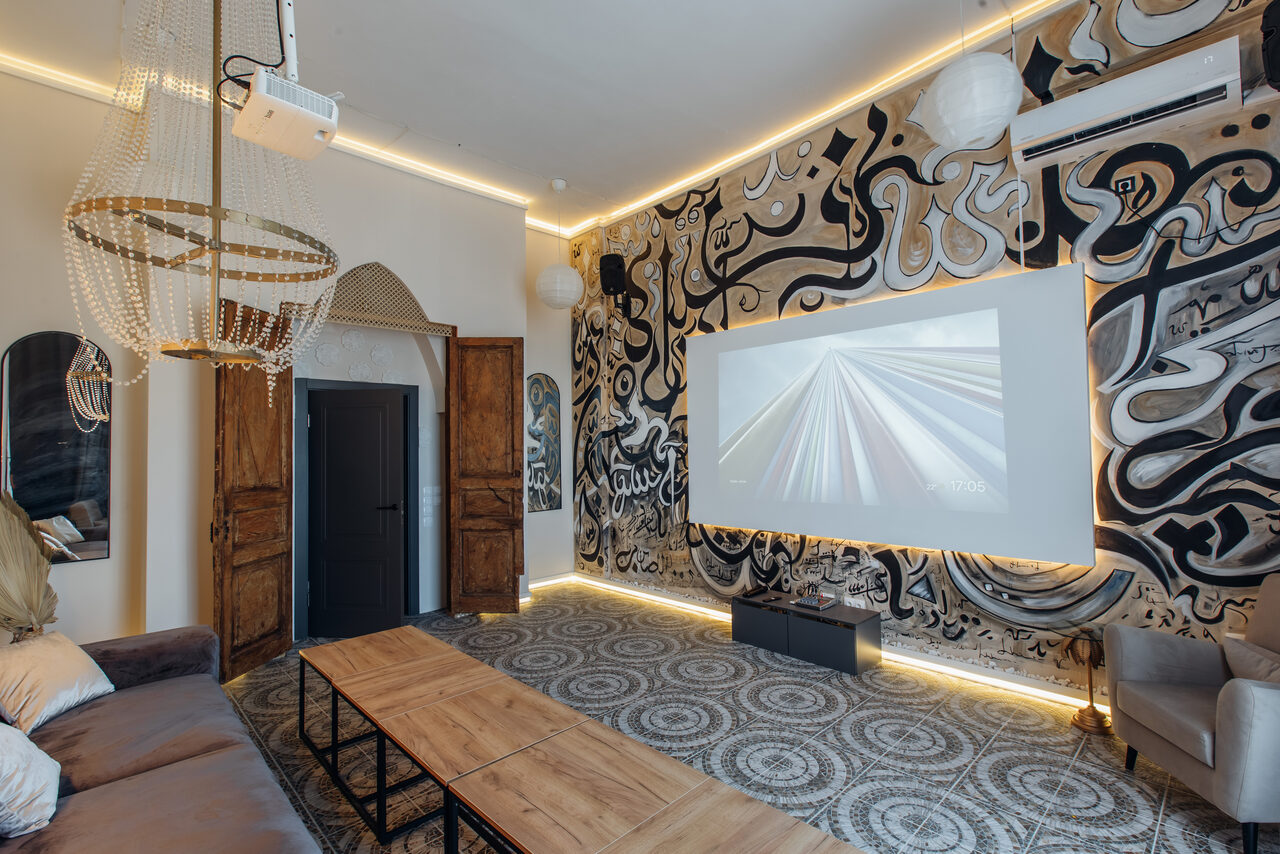 Арендовать Rubin Loft (Рубин Лофт). Зал Дубай. Гостиная с проектором, экраном и современной настенной росписью. Деревянные столы, мягкие диваны и кресла. Подвесное освещение с декоративной люстрой. Узорный пол и открытая кладка стен. Потолочный кондиционер.