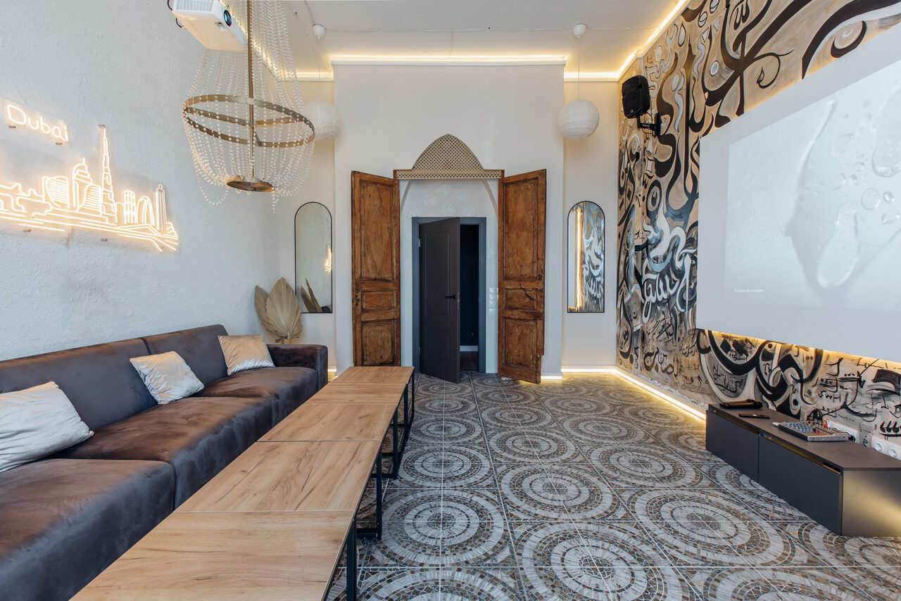 Арендовать Rubin Loft (Рубин Лофт). Зал Дубай. Гостиная с угловым диваном и подушками, длинный деревянный стол, большой экран на стене. На стене граффити. Пол с узорчатой плиткой, деревянные двери, неоновая надпись. Светильники, встроенное освещение, арочные зеркала.