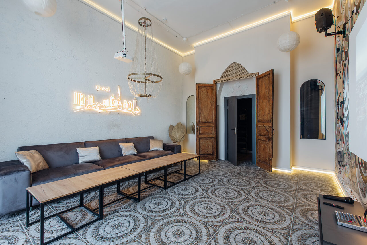 Арендовать Rubin Loft (Рубин Лофт). Зал Дубай. Современная гостиная с длинным серым диваном, деревянным столом, на стене неоновая надпись. Подвесные светильники и проектор. Пол с узорчатой плиткой, белые стены. Большие деревянные двери, зеркало, светлые фонарики, светодиодная подсветка.
