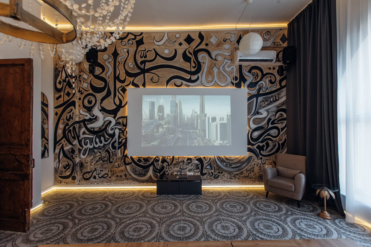 Арендовать Rubin Loft (Рубин Лофт). Зал Дубай. Лофт-пространство с граффити на стене, проектор транслирует изображение города. Темные шторы, кресло, круглый светильник, текстиль на полу с узором. Потолочная люстра, декоративные элементы из дерева и ткани.