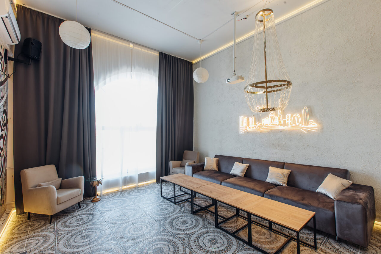 Арендовать Rubin Loft (Рубин Лофт). Зал Дубай. Просторная комната с серым диваном и светлыми подушками, длинным деревянным столом, двумя бежевыми креслами, серыми шторами, круглым подвесным светильником, декоративным неоновым освещением на стене и узорным плиточным полом.