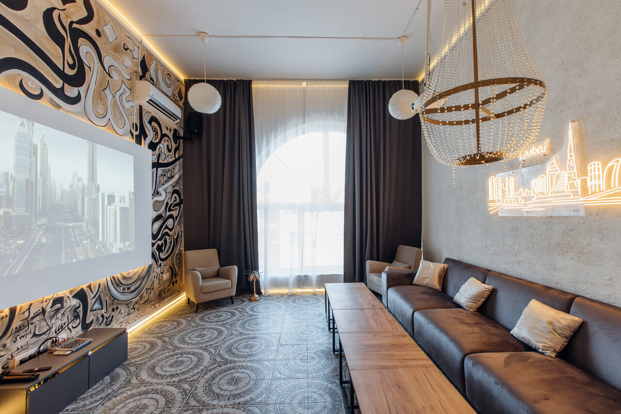 Арендовать Rubin Loft (Рубин Лофт). Зал Дубай. Комната с проектором и экраном, диваном и креслами. Стены украшены рисунками и неоновой вывеской. На полу плитка с узорами. Висит большая люстра. Есть окно с тяжелыми шторами. В помещении преобладают серые и коричневые тона.