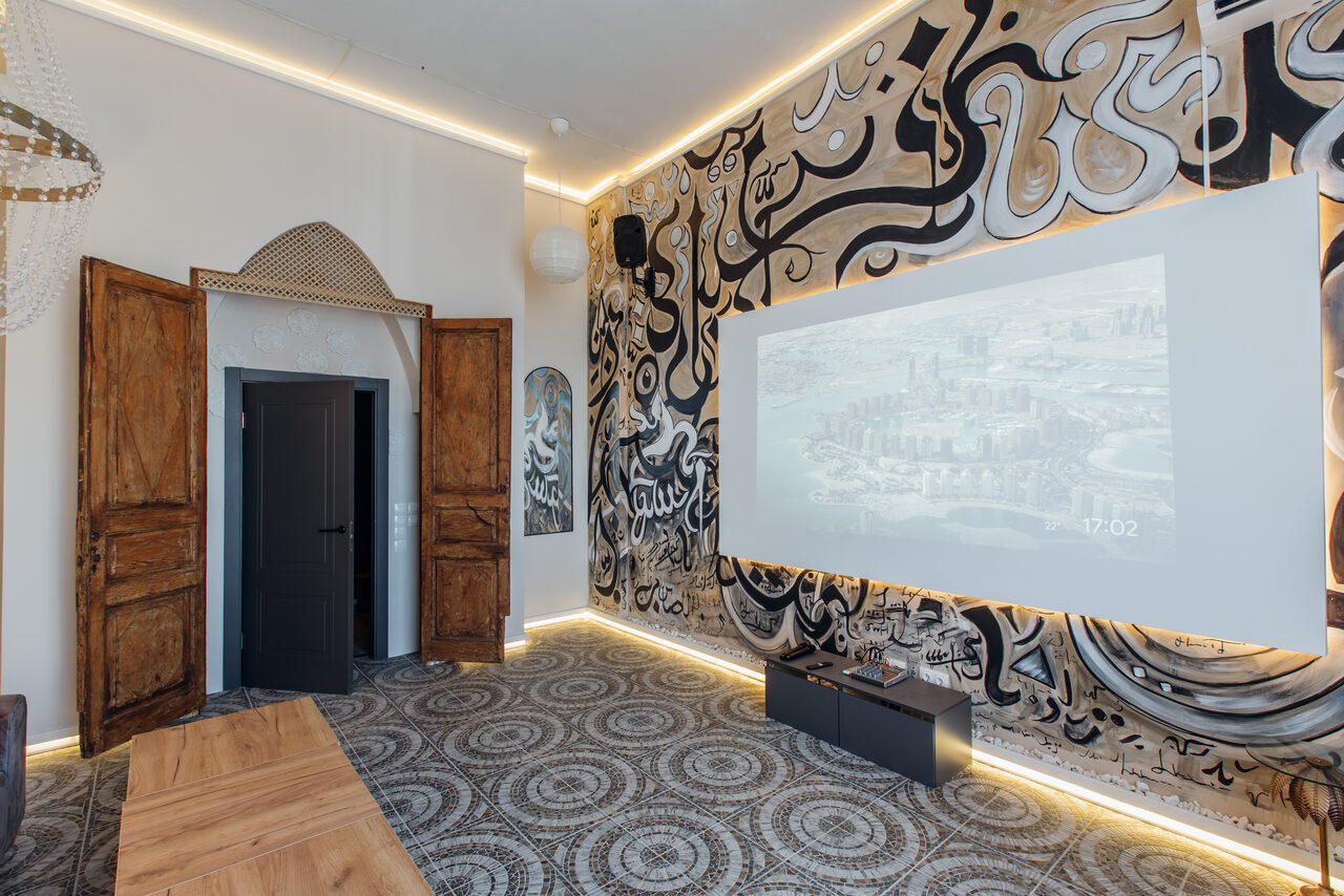 Арендовать Rubin Loft (Рубин Лофт). Зал Дубай. Пространство с высокой перегородкой и проектором. Стены украшены абстрактными черно-белыми узорами. Светодиодное освещение вдоль потолка и пола. Древесные двери и накладка. Пол выложен плиткой с узором.
