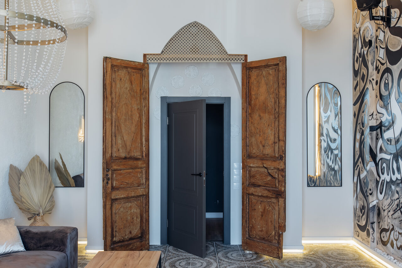 Арендовать Rubin Loft (Рубин Лофт). Зал Дубай. Аркадные деревянные двери с открытой серой дверью за ними. Белые стены, два зеркала по бокам, декоративные светильники сверху. Слева серый диван, деревянный стол. Темный узорчатый ковер на полу. Сухие листья у стены.
