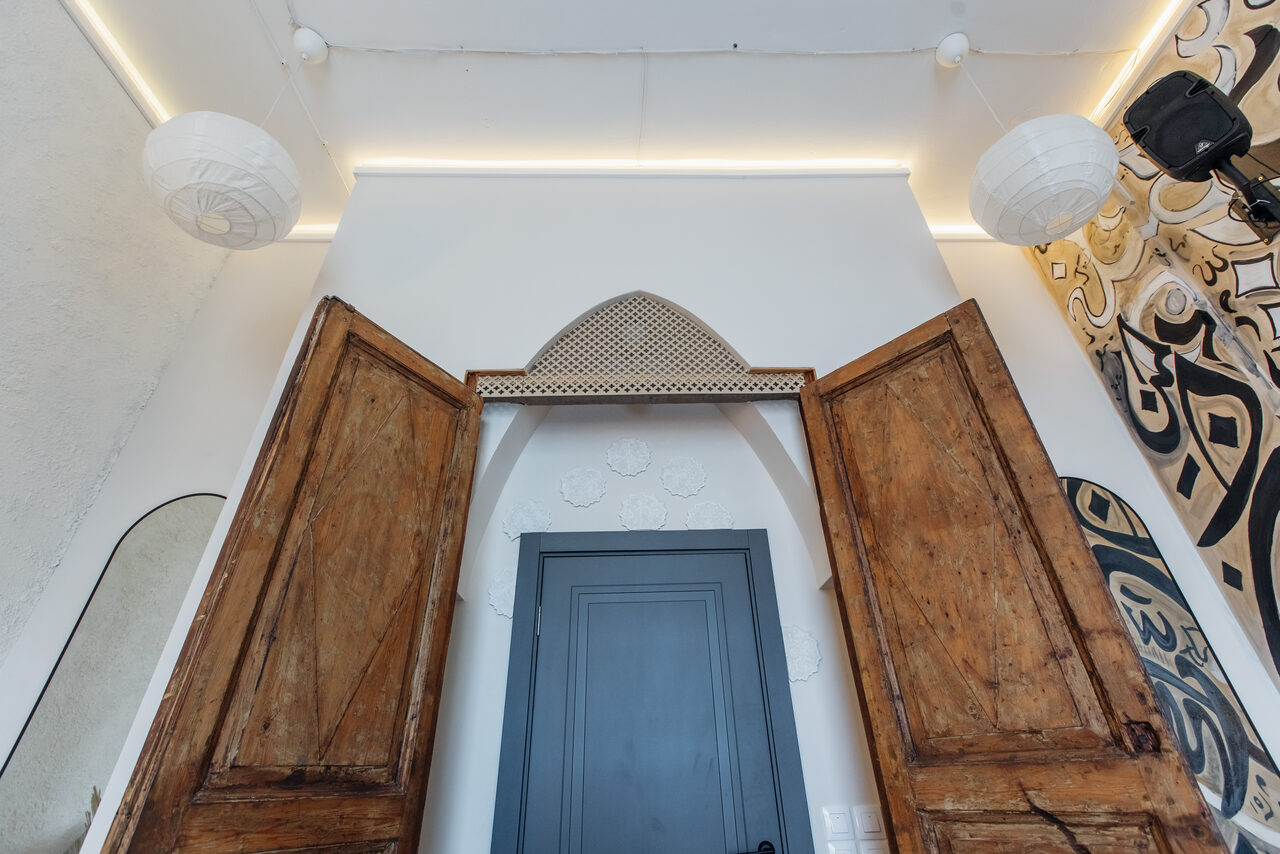 Арендовать Rubin Loft (Рубин Лофт). Зал Дубай. Интерьерное пространство с деревянными раздвижными дверями, арочным проемом, белыми стенами и декором в виде резных орнаментов. Слева зеркало, справа яркая настенная фреска. Подвесные светильники и светодиодная подсветка под потолком.