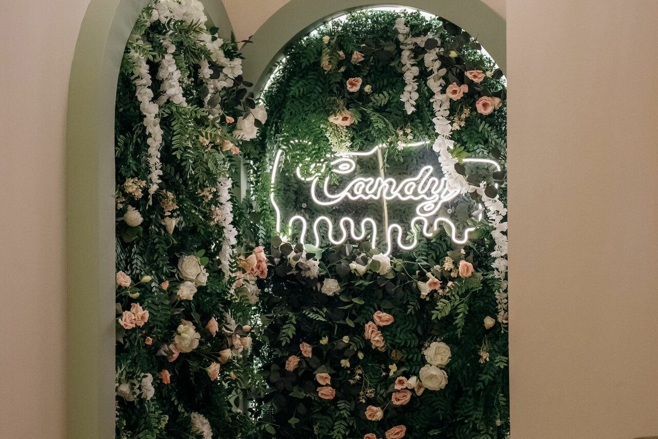 Арендовать Лофт Кэнди. Арочное декоративное оформление с неоновым белым надписью "Candy", окруженное зелеными листьями и цветами, включая розовые и белые розы. Вертикальная композиция из растений, освещение мягкое, теплые тона.