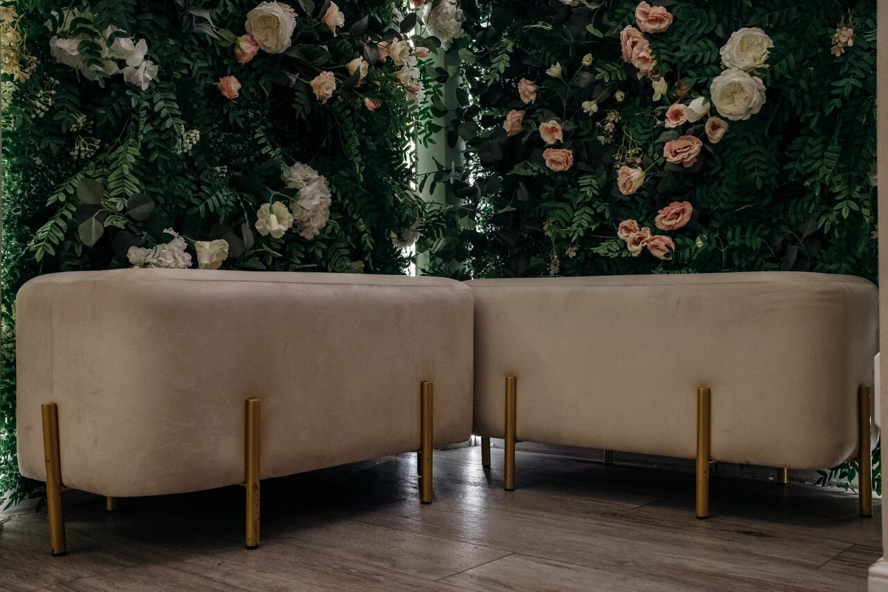 Арендовать Лофт Кэнди. Бежевая мебель с золотистыми ножками расположена на деревянном полу. На заднем плане стена украшена густой зеленью с крупными белыми и розовыми цветами. Освещение приглушенное.