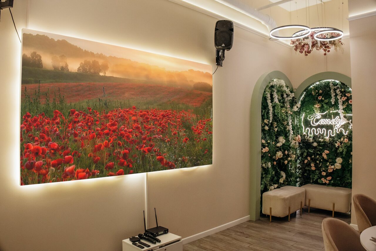 Арендовать Лофт Кэнди. Современное помещение с белыми стенами и деревянным полом. На стене большое светящееся изображение цветочного поля. В углу арка с зелеными и белыми цветами, неоновая вывеска. Мягкие сиденья бежевого цвета, круговые светильники наверху.