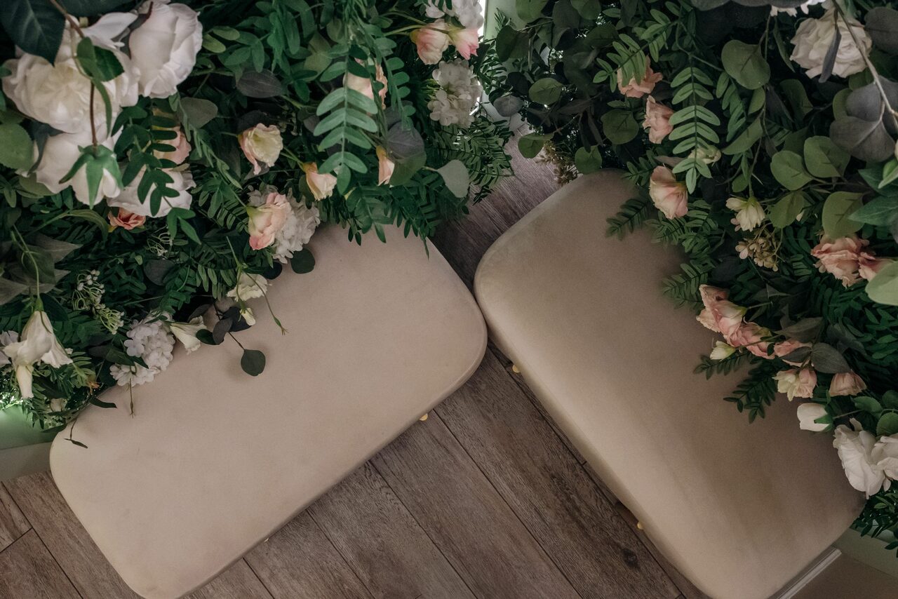 Арендовать Лофт Кэнди. Два светлых стула с мягкой обивкой, сверху обрамлены зелёными листьями и цветами в пастельных тонах. Пол из светлого дерева.