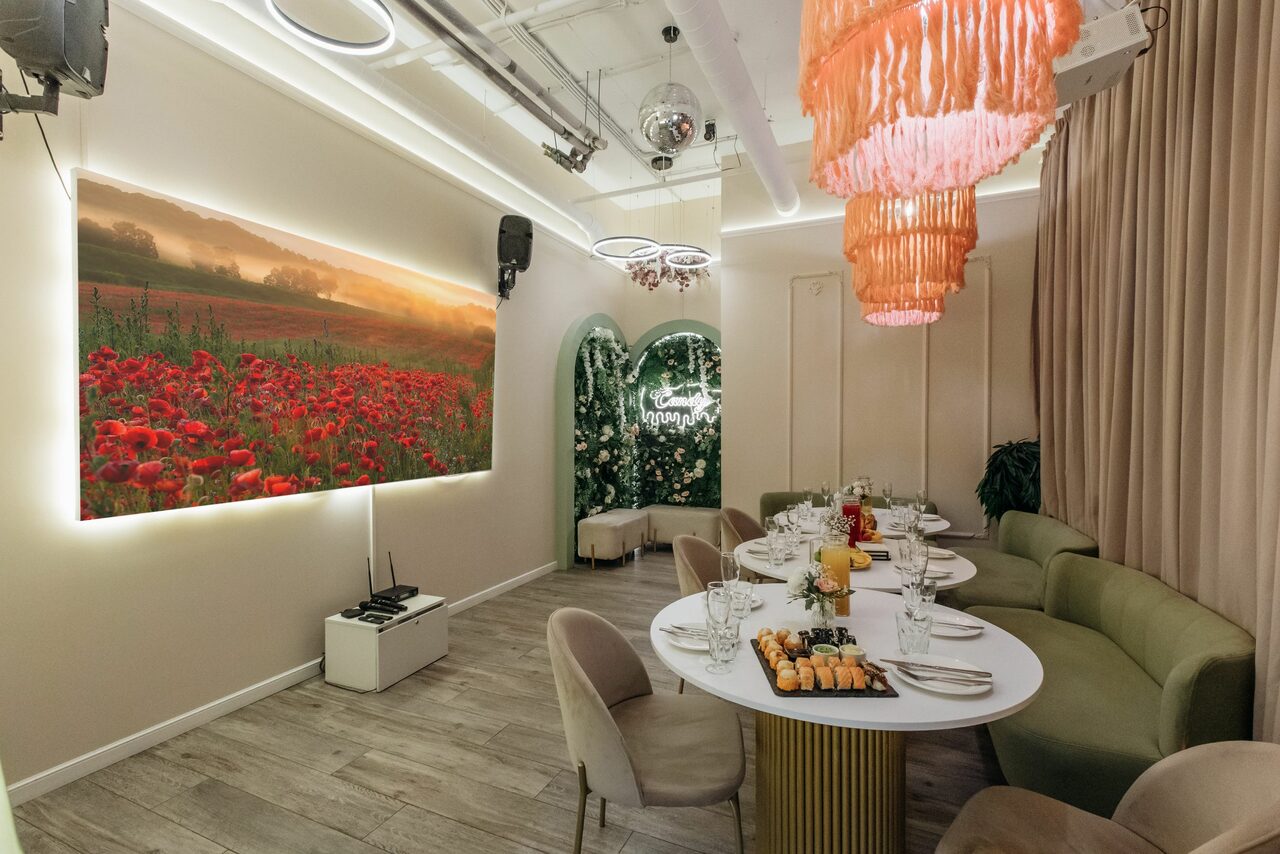 Арендовать Лофт Кэнди. Ресторанный зал с круглым белым столом и мягкими стульями; на стене большой пейзаж с полем маков и декоративный арочный элемент с цветами; потолочные лампы с оранжевыми элементами; затененные занавески и декоративное освещение.