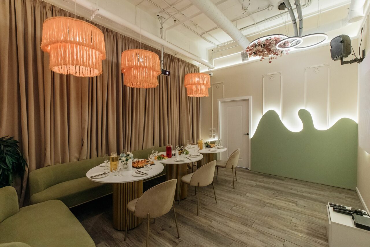 Арендовать Лофт Кэнди. Современное пространство с длинным обеденным столом и стульями, зелёные и бежевые цвета; потолочные украшения и розовые лампы; деревянное напольное покрытие; стена с волнообразным декором и подсветкой; закрытые бежевые шторы; настенное устройство.