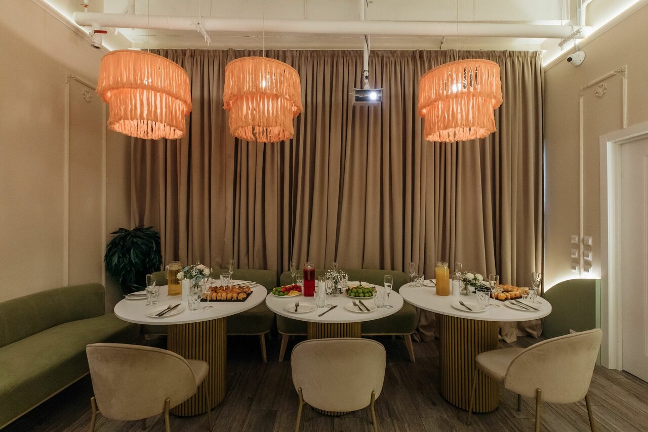 Арендовать Лофт Кэнди. Лофт со светлыми стенами и тремя круглым столами, сервированными для еды. Три подвесные светильника с бахромой, зелёный диван вдоль стены, шесть стульев, занавески и деревянный пол. Блюда и напитки на столах.