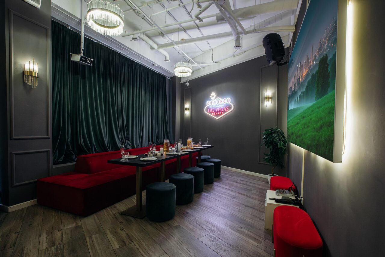 Арендовать Rubin Loft (Рубин Лофт). Лофт Вегас. Караоке-комната с темными стенами, длинный стол с черными пуфами, красные диваны, неоновая вывеска, шторы, подвесные светильники, большая картина с подсветкой, мягкое общее освещение, деревянный пол, декоративные растения.