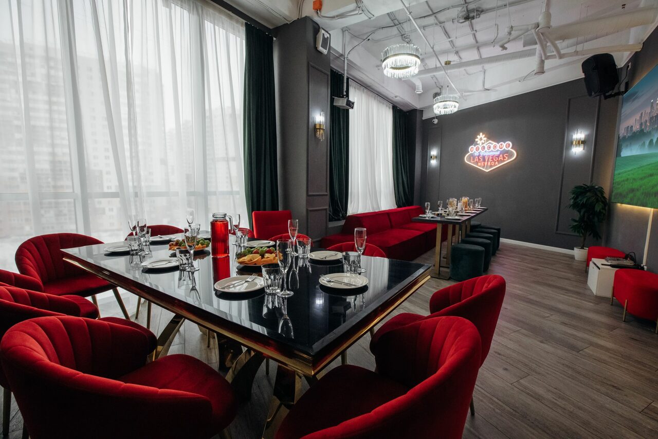 Арендовать Rubin Loft (Рубин Лофт). Лофт Вегас. Просторное помещение с большими окнами, затемнёнными шторами. Черный прямоугольный стол с красными стульями, сервирован посудой. Красный диван вдоль стены. Неоновая надпись на темной стене. Светлое деревянное напольное покрытие. Потолочные светильники и видимые коммуникации.