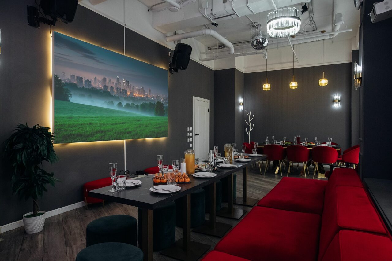 Арендовать Rubin Loft (Рубин Лофт). Лофт Вегас. Просторное помещение с серыми стенами, столами из темного дерева, красными мягкими диванами и стульями. Подвесные светильники и потолочные светильники. Крупная картина с подсветкой на стене. Посуду и бокалы расставлены на столах.