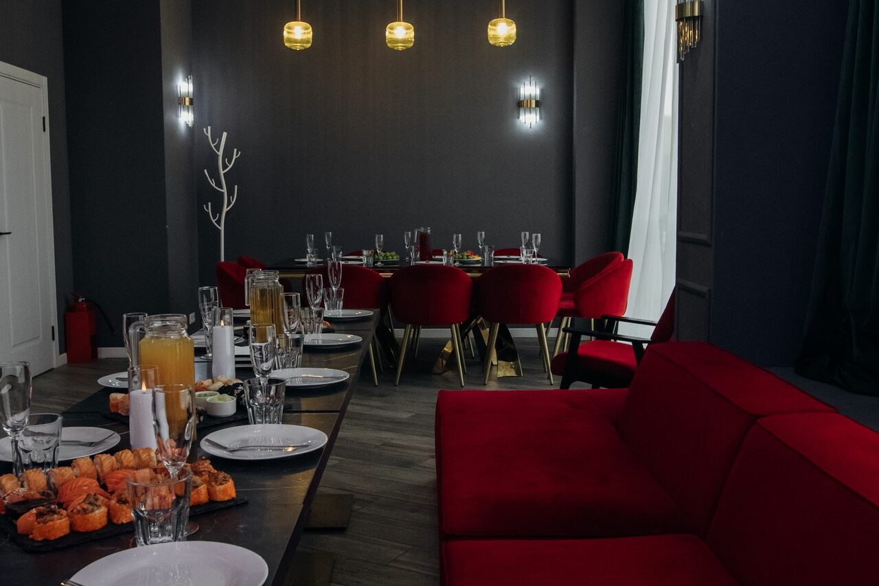 Арендовать Rubin Loft (Рубин Лофт). Лофт Вегас. Столовая с красными стульями и диваном, деревянный пол, темные стены, круглые столы, накрытые белыми тарелками и напитками, подвесные и настенные светильники, светлая штора, декоративное дерево, огнетушитель у стены.
