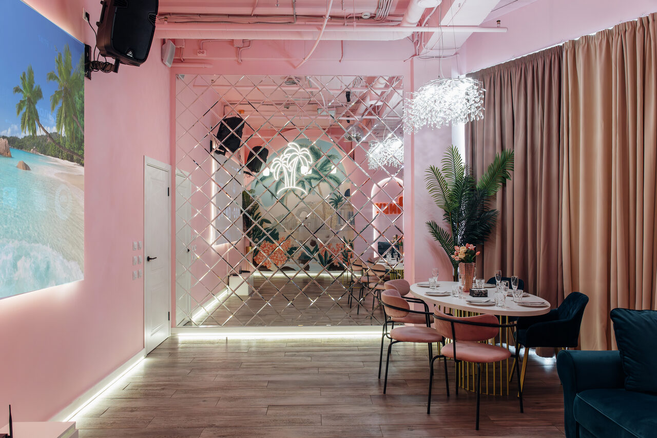 Арендовать Rubin Loft (Рубин Лофт). Лофт Пинки. Пространство с розовыми стенами, зеркальной стеной и настенным искусством с изображением пляжа. Деревянный пол, круглый стол и стулья, диван, декоративные растения, подвесные светильники. Потолок белый с открытыми коммуникациями. Фоновые шторы.