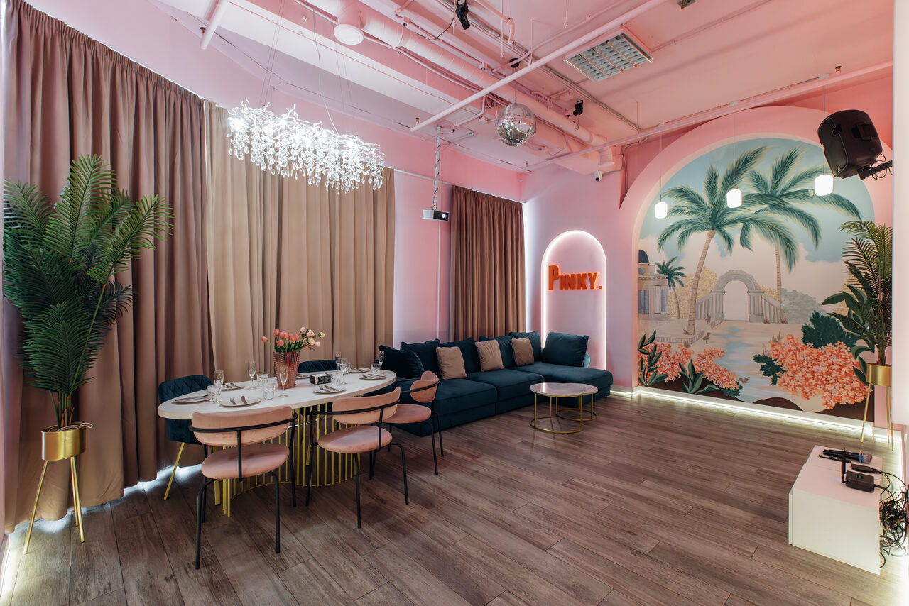 Арендовать Rubin Loft (Рубин Лофт). Лофт Пинки. Просторная комната с розовыми стенами и потолком. На стене фреска с пальмами и арками. Темный диван, круглый стол с посудой и стульями. Подвесное освещение, деревянный пол, бежевые шторы. Неоновая надпись "Phunky".