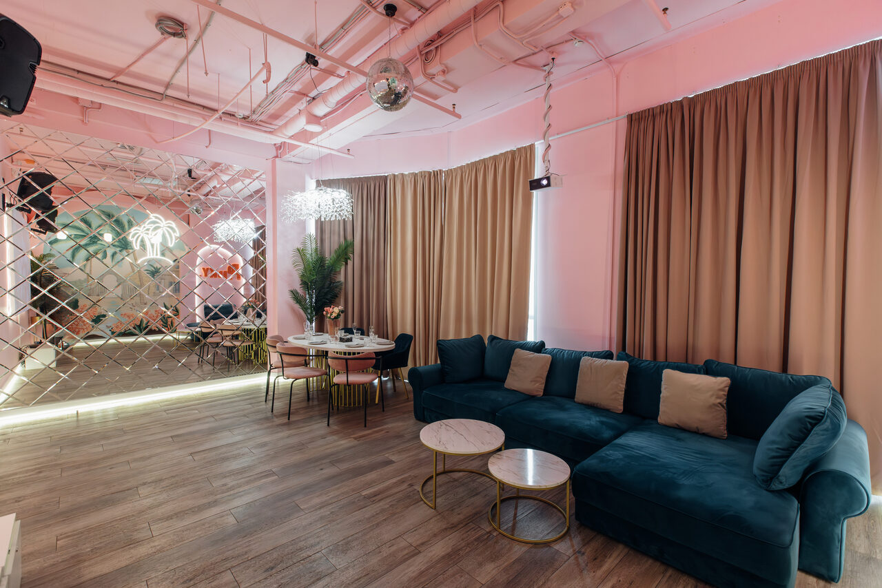 Арендовать Rubin Loft (Рубин Лофт). Лофт Пинки. Просторная гостиная с розовыми стенами и деревянным полом. Темно-зеленый диван и два круглых столика. Зеркальная стена и шторы бежевого цвета. Обеденный стол с стульями, декоративные растения, подвесной светильник. Потолок с открытыми коммуникациями.