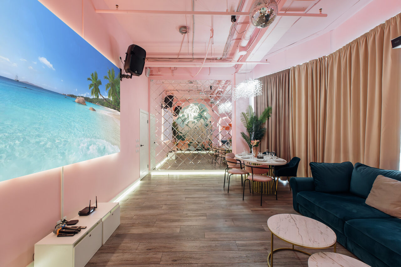 Арендовать Rubin Loft (Рубин Лофт). Лофт Пинки. Современное пространство с розовыми стенами и подвесным потолком. В комнате диван, круглый стол с мягкими стульями, на стене большой пляжный постер. На полу ламинат, на стене зеркальная плитка. Занавески, декор и несколько столиков.