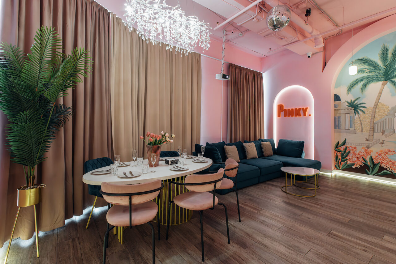 Арендовать Rubin Loft (Рубин Лофт). Лофт Пинки. Пространство с зоной отдыха и обеденной зоной. Диван и круглый стол с пятью стульями. Декоративные растения и светильники. На стене яркая роспись и текст на неоновом фоне. Пол из древесины, потолок окрашен в светлый цвет.