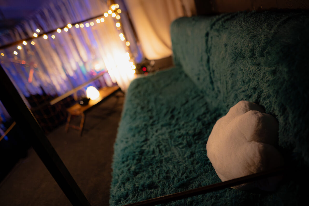 Арендовать Лес. Театральный лофт с верандой, сценой и вторым ярусом. Мягкий диван с бирюзовой обивкой, подушка в виде облака, приглушённое освещение, гирлянда с тёплыми светодиодами, белые и синие занавески, небольшой деревянный столик с круглой лампой, тёмный ковёр на полу.