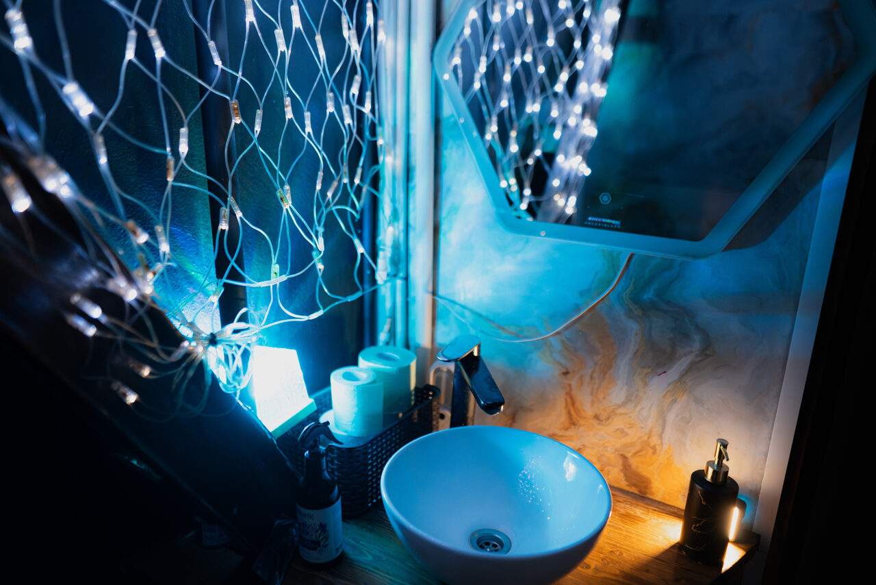 Арендовать Лес. Театральный лофт с верандой, сценой и вторым ярусом. Ванная комната с круглой раковиной на деревянной поверхности, настенным зеркалом и светодиодной гирляндой. Стены с мраморным узором, на столешнице корзина с туалетной бумагой и дозатор для мыла. Освещение холодных тонов.