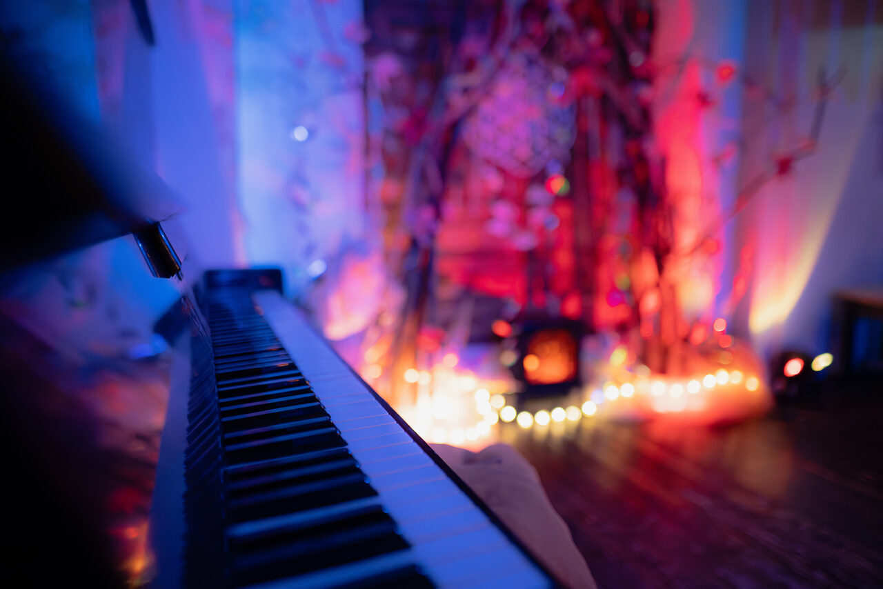 Арендовать Лес. Театральный лофт с верандой, сценой и вторым ярусом. Клавиши фортепиано слева на переднем плане. Позади него декоративная установка из веток и гирлянды с теплым освещением. Стены и пол темные, освещение разноцветное с синеватыми и красноватыми оттенками.