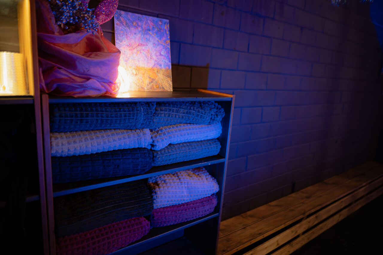 Арендовать Лес. Театральный лофт с верандой, сценой и вторым ярусом. Стеллаж с аккуратно уложенными разноцветными пледами расположен у кирпичной стены. На стеллаже картонная коробка с красочной текстурой и розовая ткань. Освещение синее и тёплое. Справа деревянная скамейка.