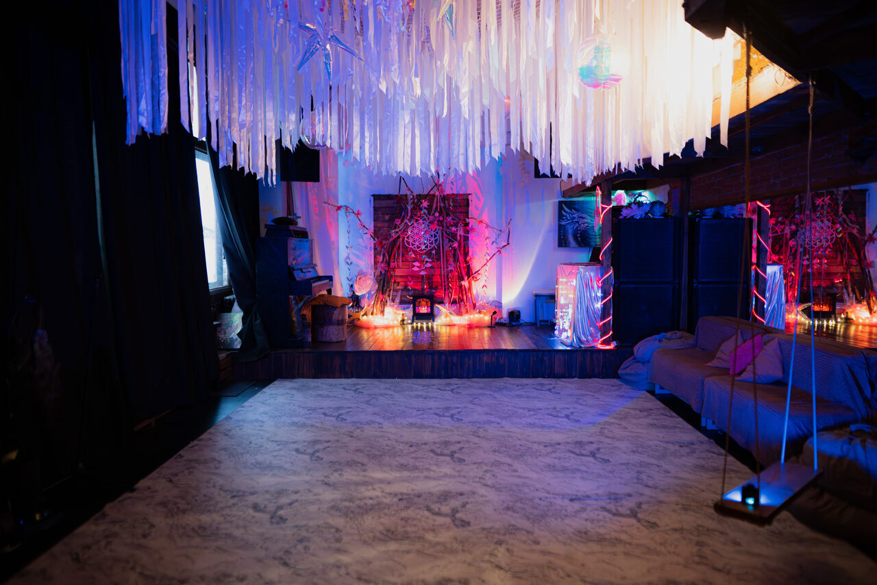 Арендовать Лес. Театральный лофт с верандой, сценой и вторым ярусом. Темное помещение с зоной отдыха. Конструкции и декор на потолке из светлых полос, мягкие диваны вдоль стены, яркая разноцветная подсветка. На возвышении декор с лампочками, стены кирпичные. Текстиль и элементы интерьера темных оттенков.