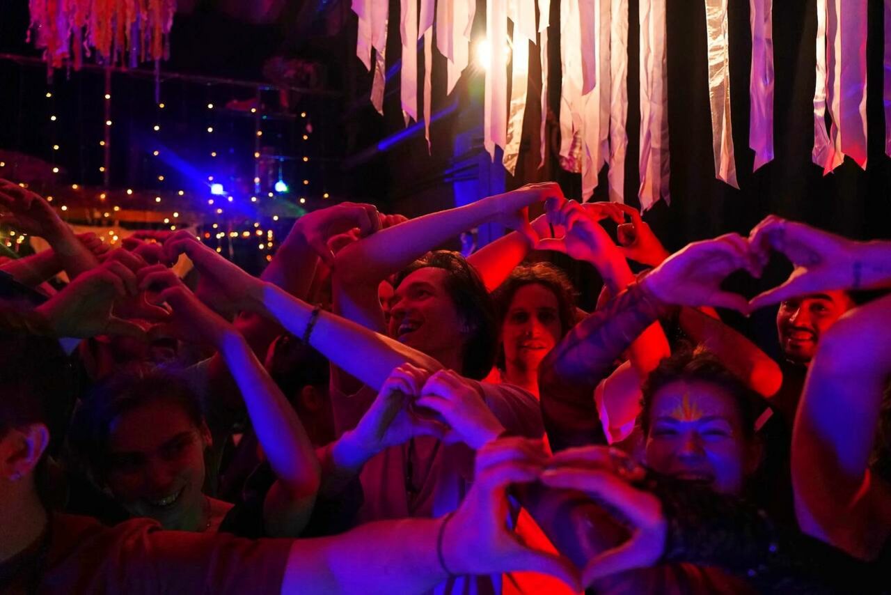 Арендовать Лес. Театральный лофт с верандой, сценой и вторым ярусом. Темное помещение с ярким разноцветным освещением. Группа людей делает жесты руками. На заднем плане свисающие ленты и светящиеся гирлянды. Теплые красные и фиолетовые тона.