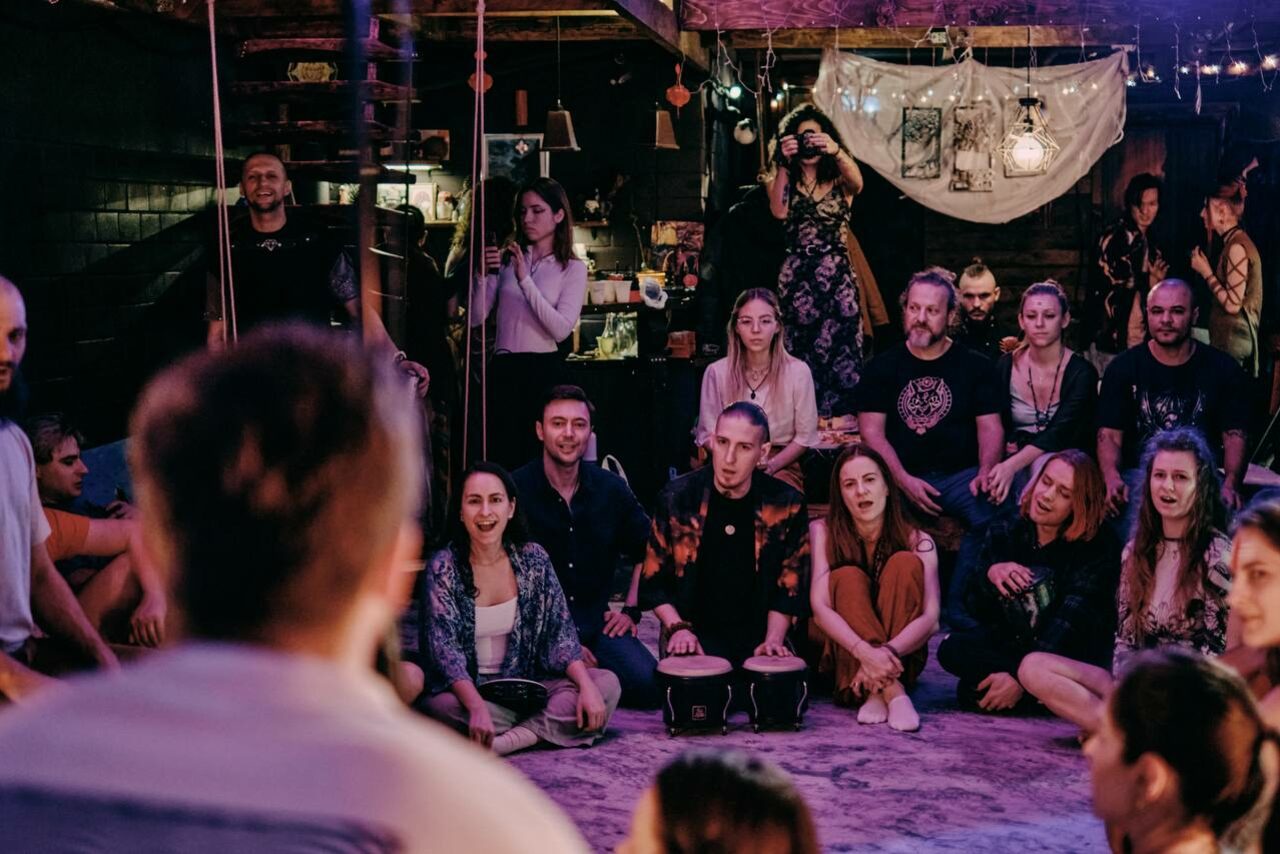 Арендовать Лес. Театральный лофт с верандой, сценой и вторым ярусом. Многофункциональное пространство с группой людей, сидящих на ковре и стульях. Настенное покрытие из тёмных кирпичей, подвесные светильники. В углу барная стойка с напитками. На заднем плане ткань с рисунками и развешенные лампы. Деревянная лестница.