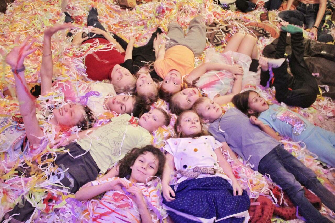 Арендовать Лес. Театральный лофт с верандой, сценой и вторым ярусом. В помещении на полу лежат дети, окружённые разноцветной мишурой. Они расположены плотно друг к другу. Несколько ярких цветов одежды и разнообразие светлых и тёмных оттенков у мишуры.