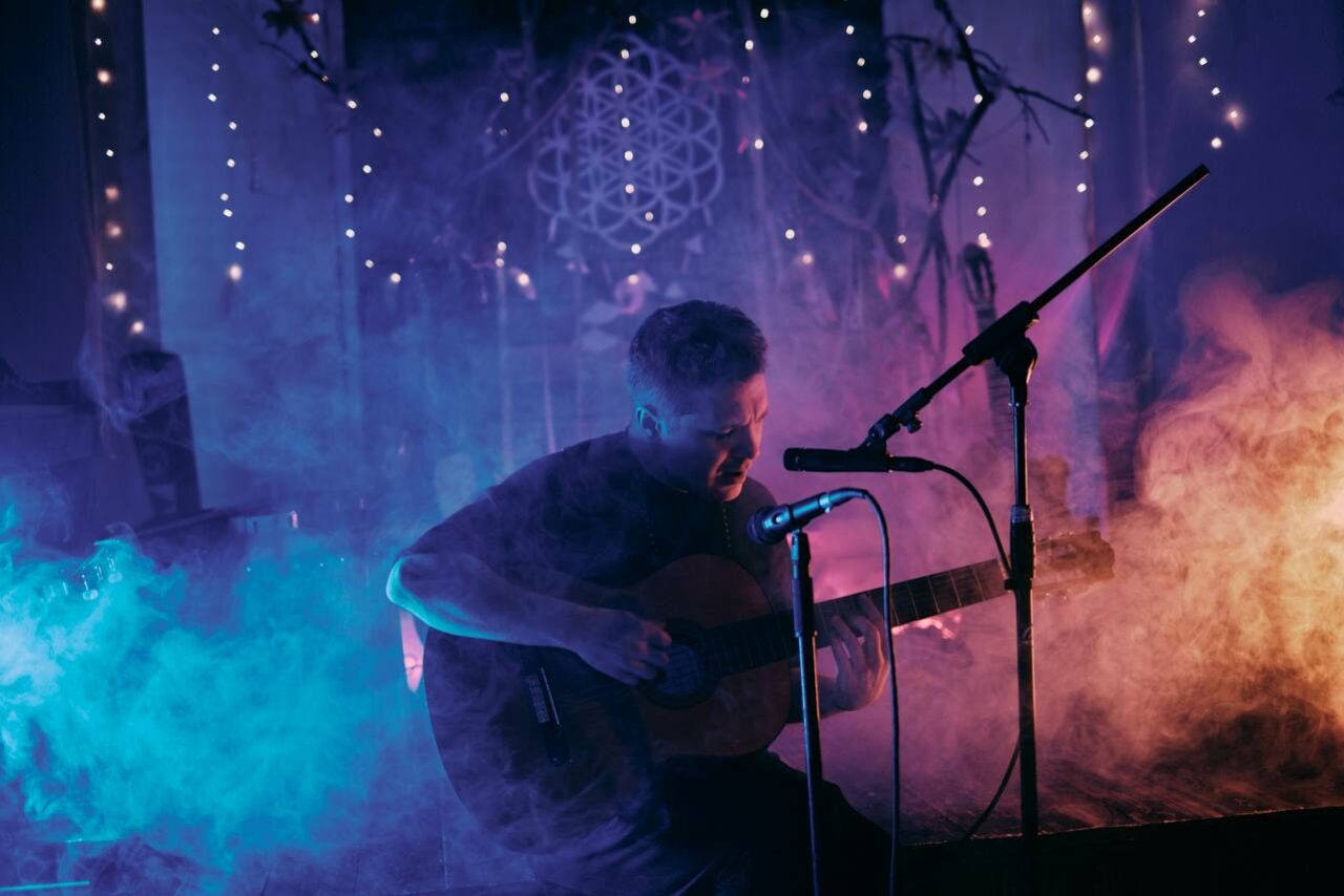 Арендовать Лес. Театральный лофт с верандой, сценой и вторым ярусом. Человек с гитарой сидит перед микрофоном в дыме, рядом стойка. На заднем плане висят небольшие огоньки. Стены украшены ветвями и иллюминацией. Цветовое освещение мягкое, в синих и фиолетовых тонах.