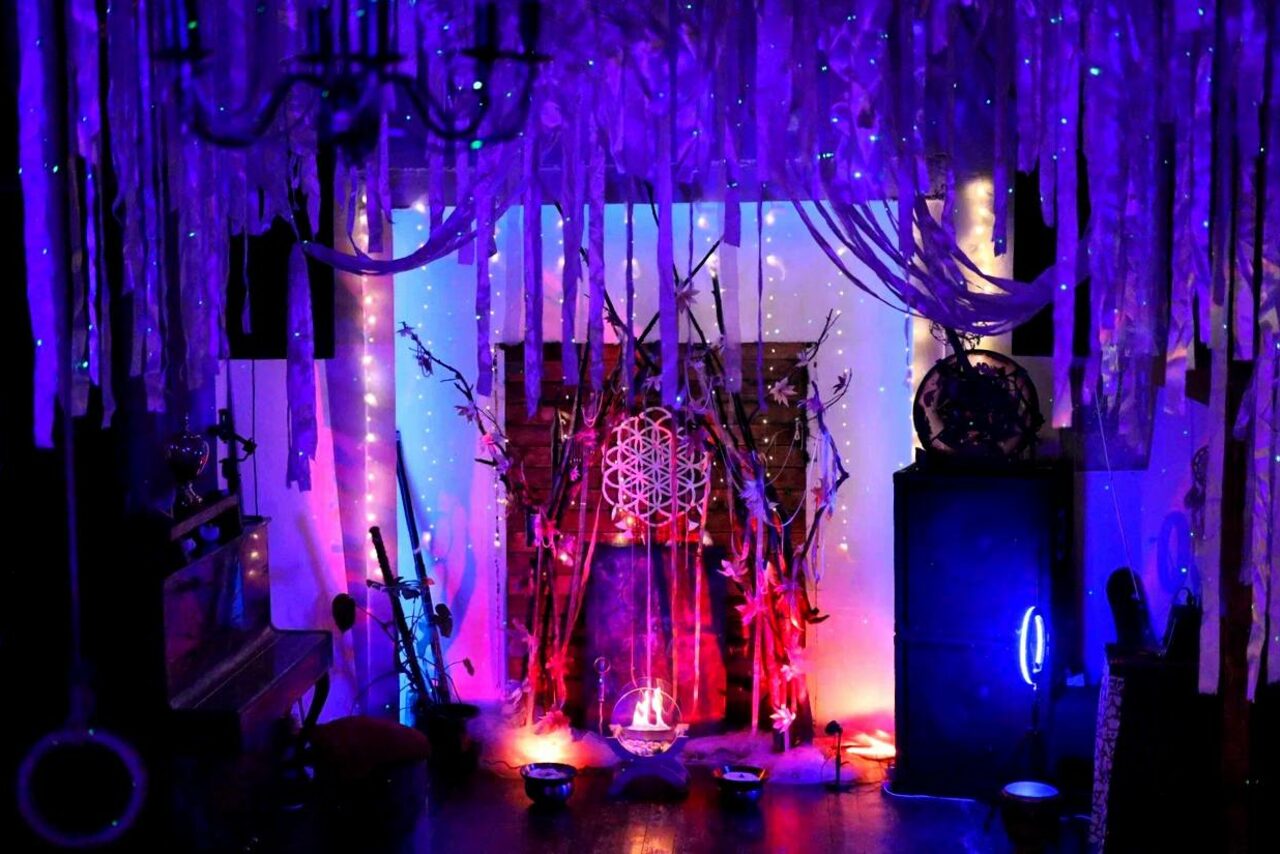 Арендовать Лес. Театральный лофт с верандой, сценой и вторым ярусом. Темное пространство с яркой подсветкой; гирлянды светодиодов в фиолетовых и голубых тонах. На полу низкие мисочки, по центру узор внутри деревянной конструкции. Потолок декорирован свисающими тканями, справа круглое зеркало, слева пианино.