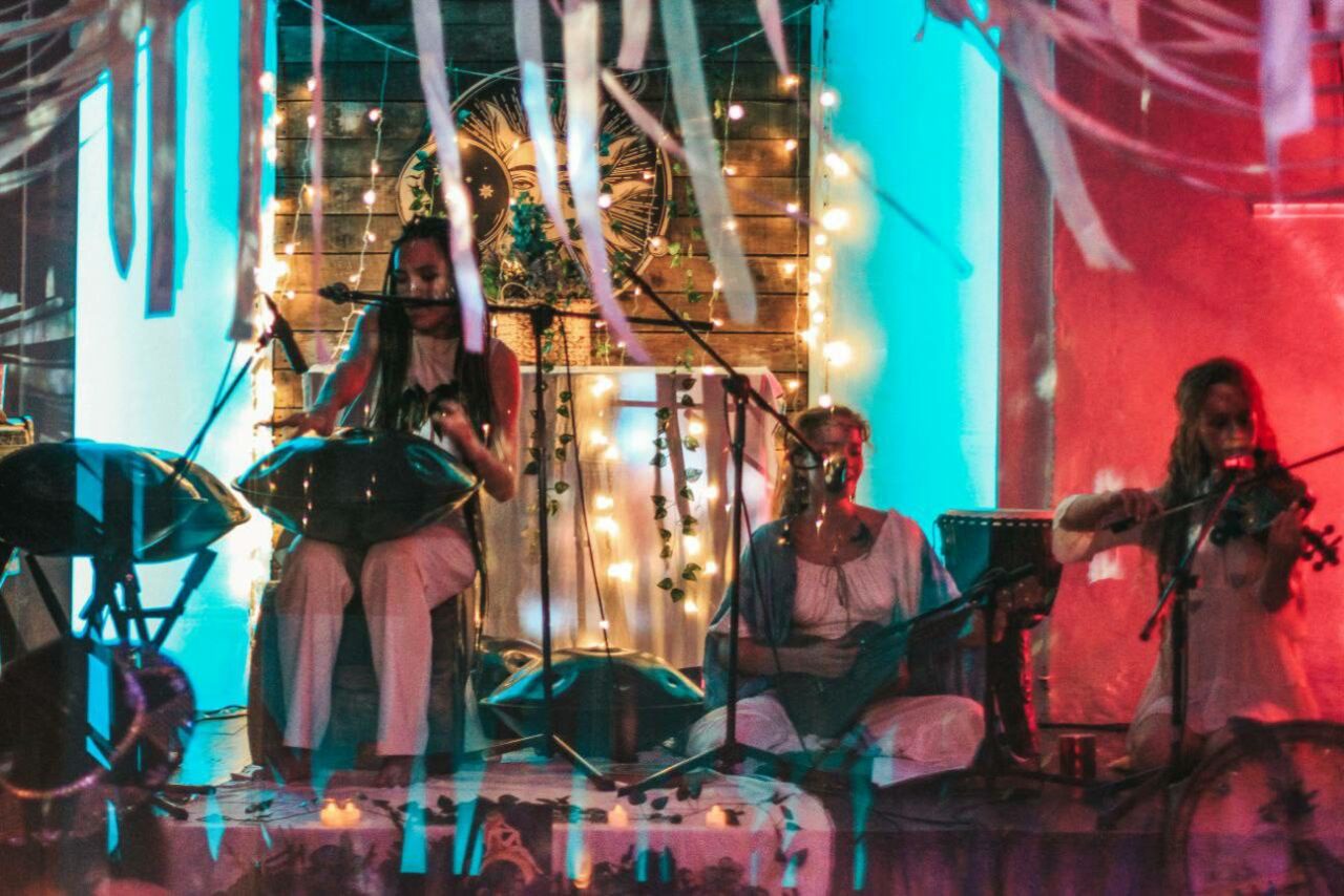 Арендовать Лес. Театральный лофт с верандой, сценой и вторым ярусом. Сцена с тремя музыкантами, деревянная задняя панель, украшенная гирляндами из лампочек, растениями и орнаментом. Микрофоны и музыкальные инструменты, приглушенное освещение в синих и розовых тонах, полупрозрачные ленты на переднем плане.