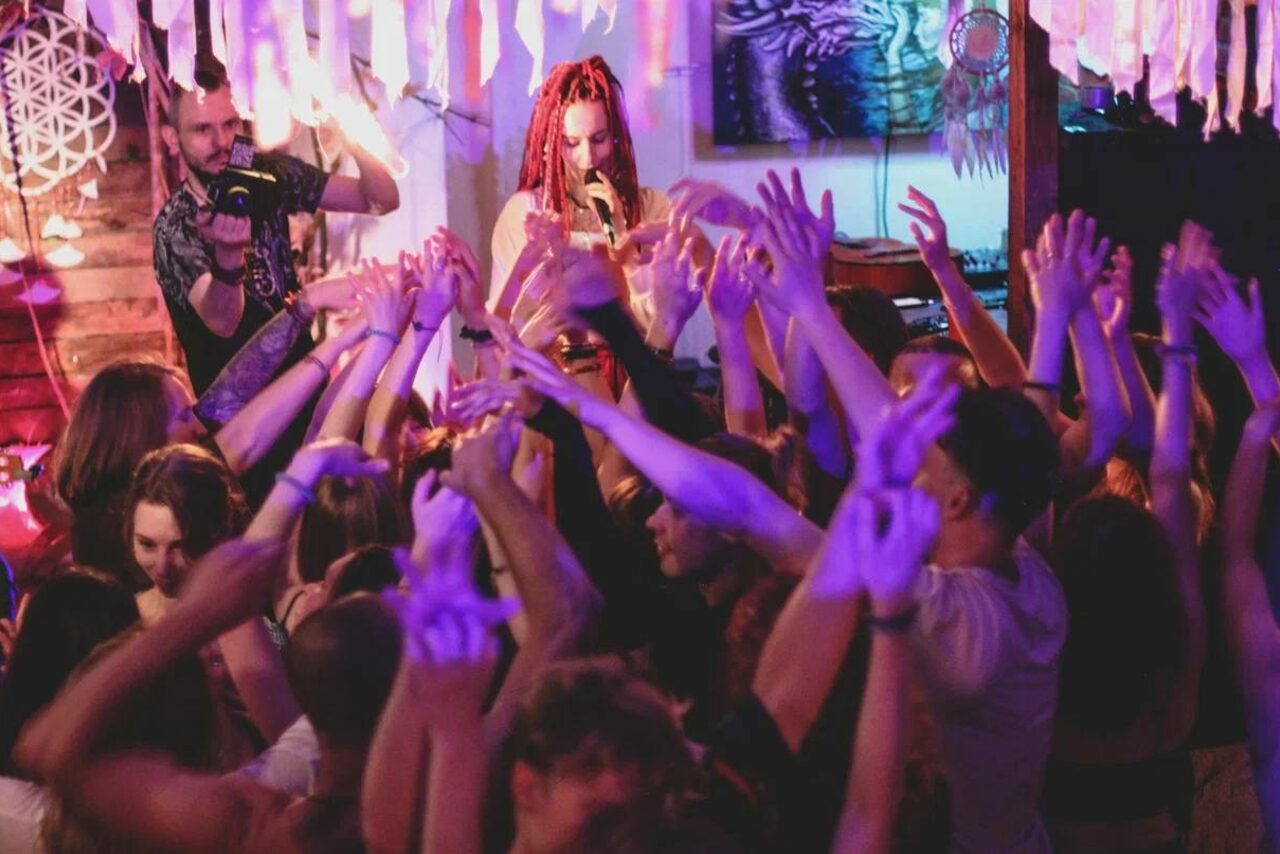 Арендовать Лес. Театральный лофт с верандой, сценой и вторым ярусом. Пространство с высокой плотностью людей, сцена с музыкантом и оператором, яркое освещение с фиолетовыми и розовыми оттенками, декор в виде бумажных лент, деревянные элементы, стена с фотографией и декором, диджейский пульт.