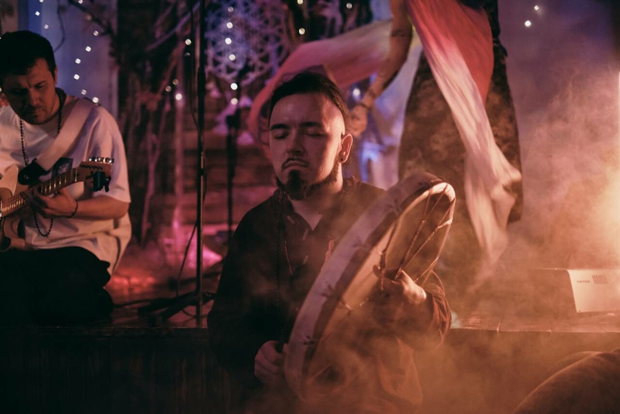 Арендовать Лес. Театральный лофт с верандой, сценой и вторым ярусом. Темное помещение с приглушенным освещением. В центре мужчина играет на ударном инструменте. Рядом человек с гитарой. Задний план украшен светящимися гирляндами и тканями. Атмосфера дымчатая.