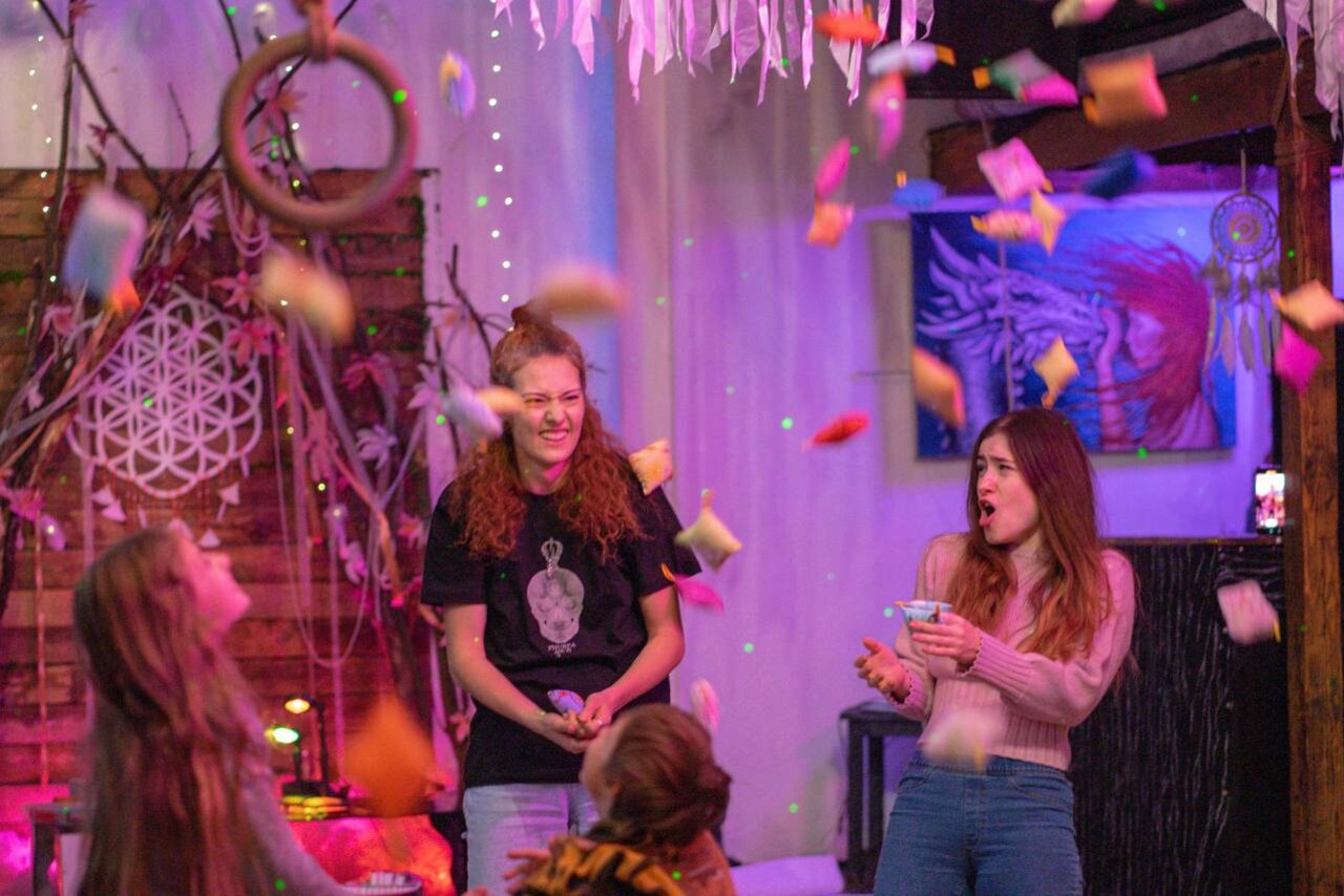 Арендовать Лес. Театральный лофт с верандой, сценой и вторым ярусом. Креативное пространство с деревянными стенами и декоративными элементами. Потолочное украшение из бумаги. Яркое разноцветное освещение. Люди с подушками. Картина на стене. Декор из натуральных материалов, гирлянды. Цвета: коричневый, фиолетовый, белый.